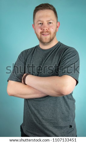 Bearded casual young man in a grey t-shirt looking pensively at the camera with his with crossed arms iup frontal studio portrait