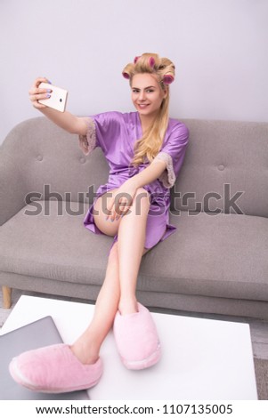 Blond woman having fun taking pictures with curlers in her hair.Beautiful attractive female wearing lilac pajamas and pink furry slippers taking selfies in living room on sofa.