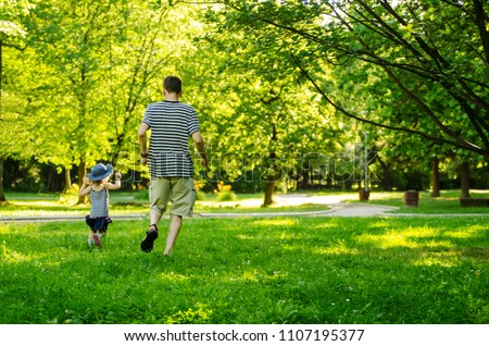 Father and toddler daughter running through the green grass.Back of dad and little child chasing in park.Copy space  Royalty-Free Stock Photo #1107195377