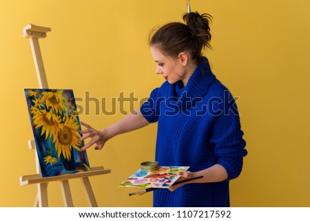 Girl artist paints sunflowers oil paints on canvas. She is wearing blue sweater. Woman is holding brush and palette with paints. She puts paint on her fingers.