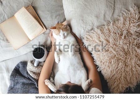 Cute ginger cat is sleeping in the bed on warm blanket. Cold autumn or winter weekend while reading a book and drinking warm coffee or tea. Hygge concept. Text on the pages is not recognizable. Royalty-Free Stock Photo #1107255479