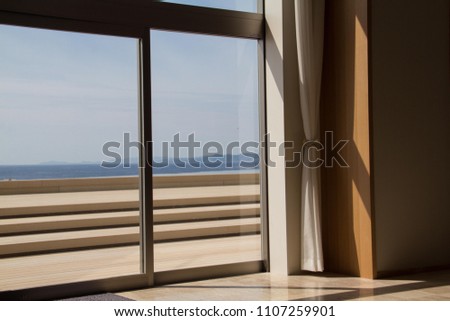 A simple modern window, with plain white curtain, and a view to a summer sunlit modern cream colored veranda and deck overlooking the sea. Sunlight streaming into the building. Royalty-Free Stock Photo #1107259901