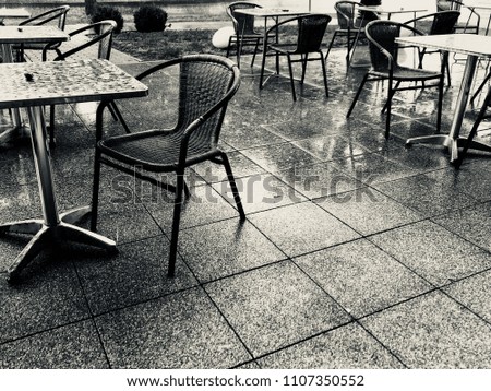 A rain shower on a summer day. View to the terrace of a coffee shop. Close up of empty tables, chairs, patio slabs with light reflections. All wet with visible raindrops. Black and white photo.