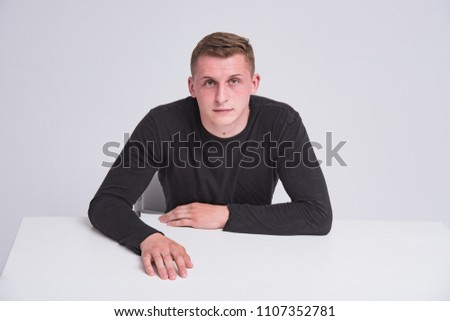 portrait of a cute young guy on a white background sitting at a table in different poses with different emotions. He sits right in front of the camera and looks happy