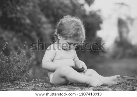 Happy kid having fun. One small curious baby boy with blonde curly hair holding and playing on black mobile phone sitting on ground outdoor looking down on natural background, horizontal picture