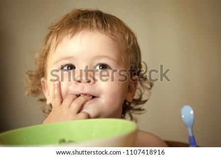 Happy kid having fun. Portrait of sweet small smiling baby boy with blonde curly hair and round cheecks eating from green plate holding spoon and lick fingers closeup, horizontal picture