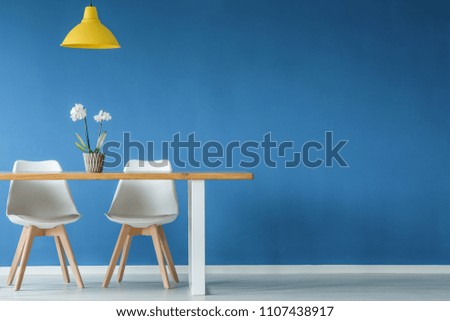 Two chairs, flower, yellow lamp and wooden table on a blue, empty wall in dining room interior