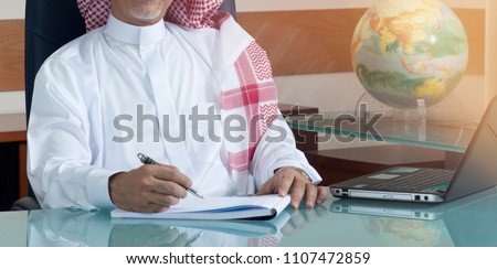 Senior Saudi Businessman Hand Writing At His Desk with Laptop Royalty-Free Stock Photo #1107472859
