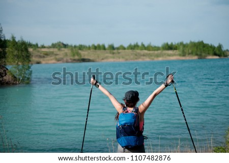 Photo from back of female tourist with arms raised with walking sticks on lake background with summer day