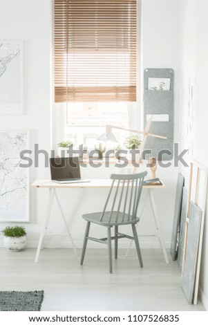 Grey wooden chair standing by the desk with lamp and laptop with empty screen in white room interior with window and fresh plants