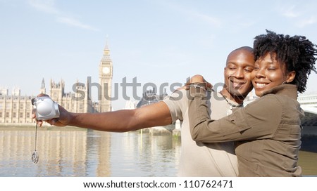 Panoramic view of a young african american couple taking pictures of themselves while on vacations in London, standing in front of Big Ben.