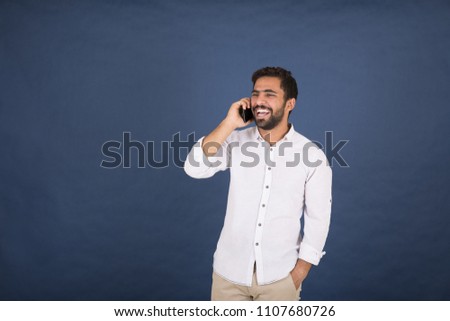 Handsome man wearing a casual outfit, holding his mobile having a phone call with his friend, and laughing, standing on a navy background.