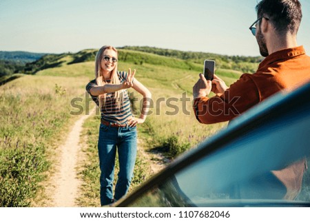 man taking picture of girlfriend doing peace sign near car on rural meadow 