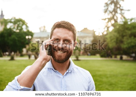 Happy photo of smiling adult man 40s in white shirt looking at you while talking on black smartphone during walk in green park
