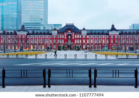The majestic historical Tokyo Train Station stands before modern buildings in gloomy bluish morning twilight and a passenger walks on the pedestrian promenade in foreground in Chiyoda, Tokyo, Japan