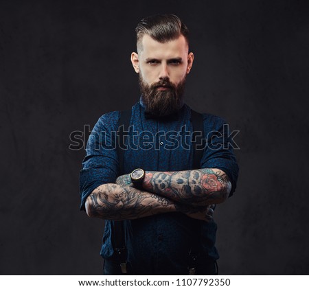 Portrait of a handsome old-fashioned hipster in a blue shirt and suspenders, standing with crossed arms in a studio.