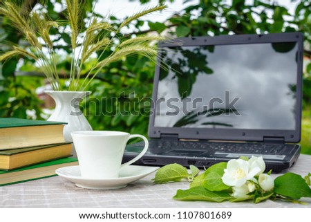 A cup with tea, a laptop, a computer, a bookshine table in the garden on a summer morning, a place to work, a concept of freelancing