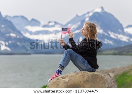 Young smiling Caucasian blond woman is sitting on rock holding American flag in her hands with Rocky Mountains and Sherburne lake background on Independence Day 4th of July in Glacier Park, Montana