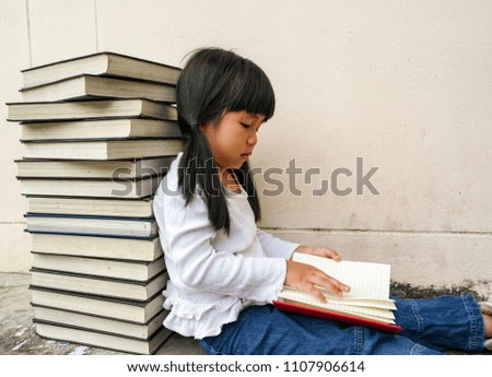 The little Asian girl is reading a book beside a pile of big books. 