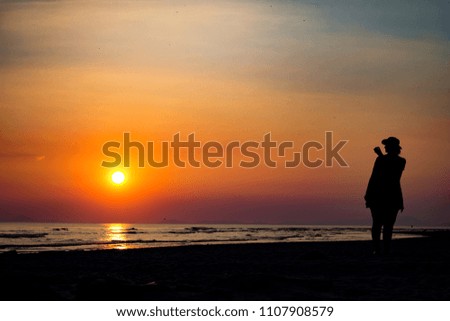 A woman stands on the beach taking a photo as the sun sets over the ocean. The sky fades into deep oranges while the foreground is black as night. 