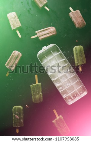 Green soothie and raspberry popsicles with an ice cube containr on a dark background, Colored lare with oposite colors green and pink.