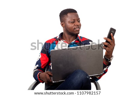 young african sitting in multicolored shirt looking at mobile phone while smiling with laptop on thighs.