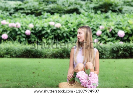 Beautiful young woman with blonde hair sitting on the grass in park, holding hands her straw bag with lilac hydrangea flowers and looking away