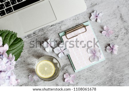 desktop flatlay items: laptop, notepad, hydrangea flower and petals, glass of water with lemon lying on grey marble background