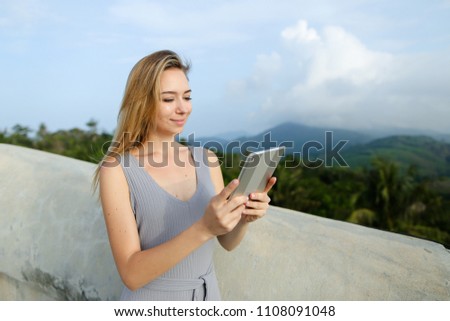 Young blonde woman using tablet in mountains background, wearing grey shirt. Concept of modern technology and nature.