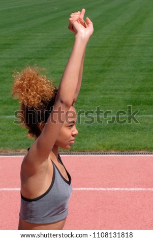 Portrait of a girl of African-American type, engaged in sports or fitness. A young woman in a gray T-shirt raised her hands up, next to the green grass of the park or stadium. Vertical picture