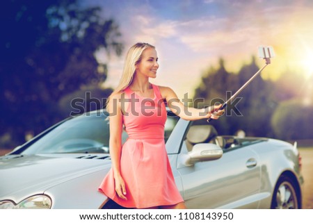 travel, road trip and people concept - happy young woman at convertible car taking picture by smartphone selfie stick over summer sunset background