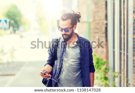 people, technology, travel and tourism - man with earphones, smartphone and bag walking along city street and listening to music 
