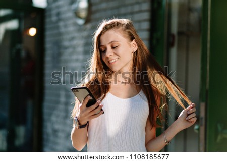 Cheerful beautiful young woman with long light-brown hairs using smartphone while walking on sunny street and plays with her hair, summer, weekend, good emotions, modern young woman