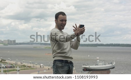 Young man makes selfie with mobile phone near the river on cloudy day