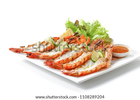 Grilled sliced black tiger prawns with baked potatoes, slice of lemons, fresh vegetables and chili sauce on white square plate, isolated on white background with shadow, low angle front side view.