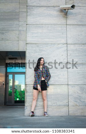 Young hipster teenager with long hair standing against the wall in the street, wearing colorful shirt in cage and black shorts on the urban background. concept of freedom purposeful teenager