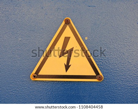 
A yellow sign of high voltage, attached by rusty screws to a dark blue wall.					