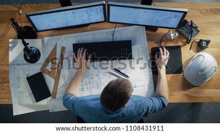 Top View of architectural Engineer Working on His Blueprints, Holding Tablet Computer, Using Desktop Computer Also. His Desk is Full of Useful Objects and Evening Sun. Royalty-Free Stock Photo #1108431911