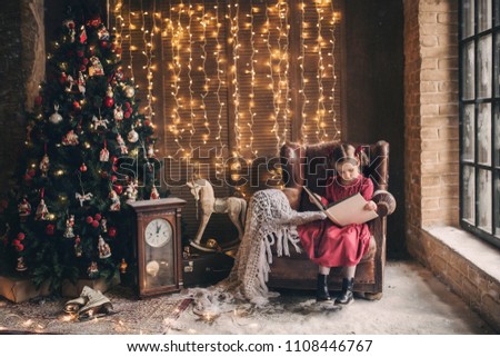 little girl is sitting in a chair and thumbs through a vintage photo album next to a Christmas tree on a background of Christmas lights, photo in retro style Royalty-Free Stock Photo #1108446767