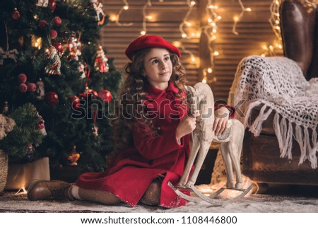 little girl in a red coat and beret hugs a vintage wooden horse next to a Christmas tree on a background of Christmas lights, photo in retro style