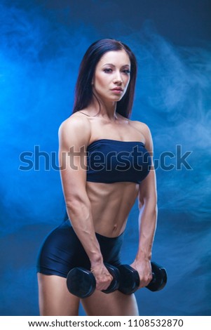 athletic young woman doing a fitness workout with dumbbells on black studio background. Around the shrouds the model smoke.