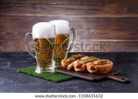 Photo of two mugs of foam beer, green grass with football, hotdogs