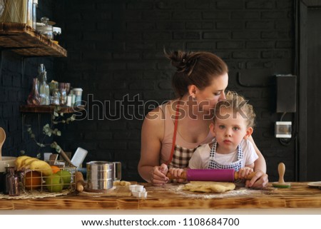 Young mother and daughter prepare cookies in kitchen. They are in aprons. Little girl rolls dough with rolling pin. Woman kisses child, encouraging diligence. Family time.