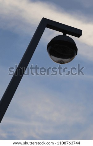 Outdoor view from bellow of a metallic lamppost. Vertical pole and round lantern with a blue grey cloudy sky in background. Abstract image of an isolated element in the space. Urban abstract picture. 