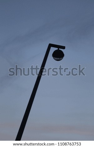 Outdoor view from bellow of a metallic lamppost. Vertical pole and round lantern with a blue grey cloudy sky in background. Abstract image of an isolated element in the space. Urban abstract picture. 