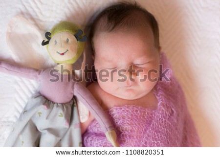 Medium horizontal frontal view of cute round-faced newborn sleeping baby girl in mauve wool wrap lying on her back on white bedspread next to a soft bee doll