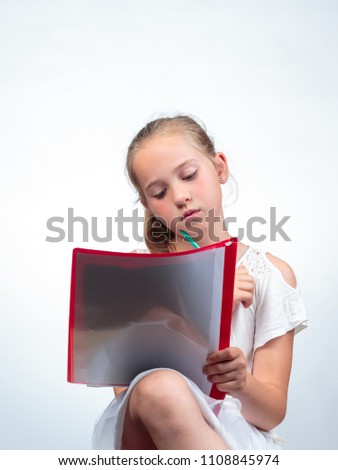 Cute 10-year-old caucasian schoolgirl looking at or reading in an note pad while holding a pencil against her chin. Against a light background