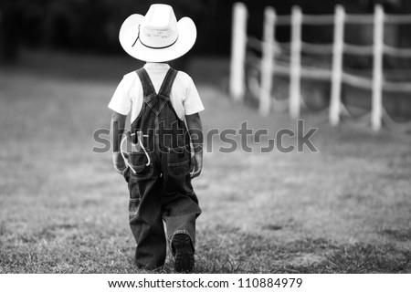 Picture of a little boy in a cowboy hat and overalls, walking away with an ole-fashioned slingshot in his back pocket.