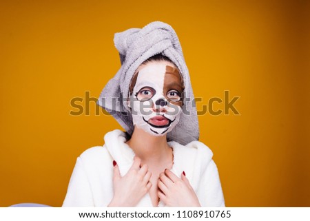 a funny young girl with a towel on her head and a white robe looks after herself, on her face a moisturizing mask with a dog's face