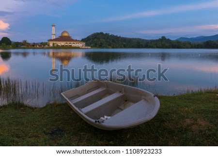 Beautiful colorful sunrise view of a mosque by a calm lake with reflection and an unknown boat in the foreground in Selangor, Malaysia.